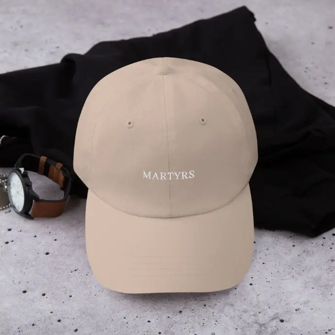 Casquette beige unisexe de la marque MARTYRS, une marque engagée qui met en lumière la culture afro-descendante à travers la mode. Le logo blanc "MARTYRS" est brodé à l’avant pour un style minimaliste et moderne. Posée sur un fond urbain en béton, accompagnée d’une montre au bracelet en cuir et d’un vêtement noir, cette casquette incarne l’alliance parfaite entre élégance, streetwear et identité culturelle.